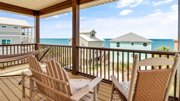 Une terrasse en bois avec deux fauteuils Adirondack et un fauteuil en osier, donnant sur une plage bordée de maisons et sur l’océan.