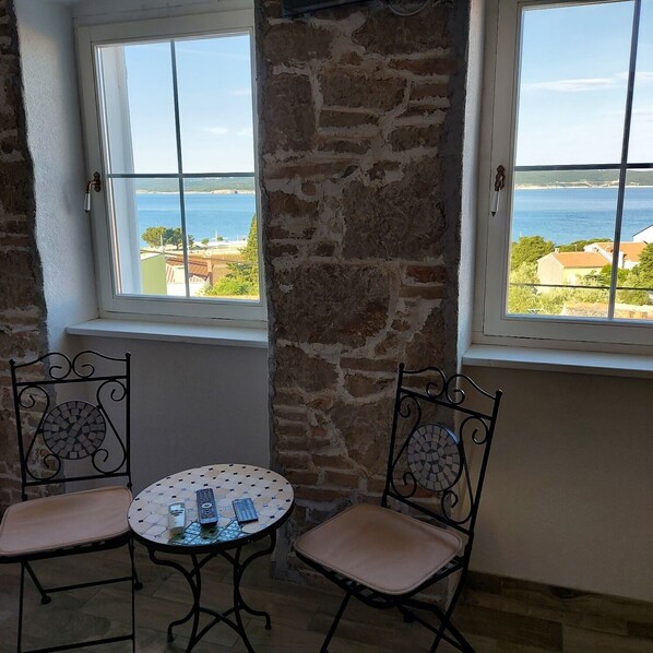A room with two windows overlooking a coastal view, a stone pillar, and a small round table with chairs.