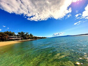 A coastal view with clear water, a beach, and a building with a balcony overlooking the sea.