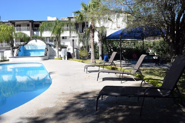 A swimming pool with lounge chairs and a covered area, surrounded by buildings and palm trees. - Hotel Virrey (Reynosa)