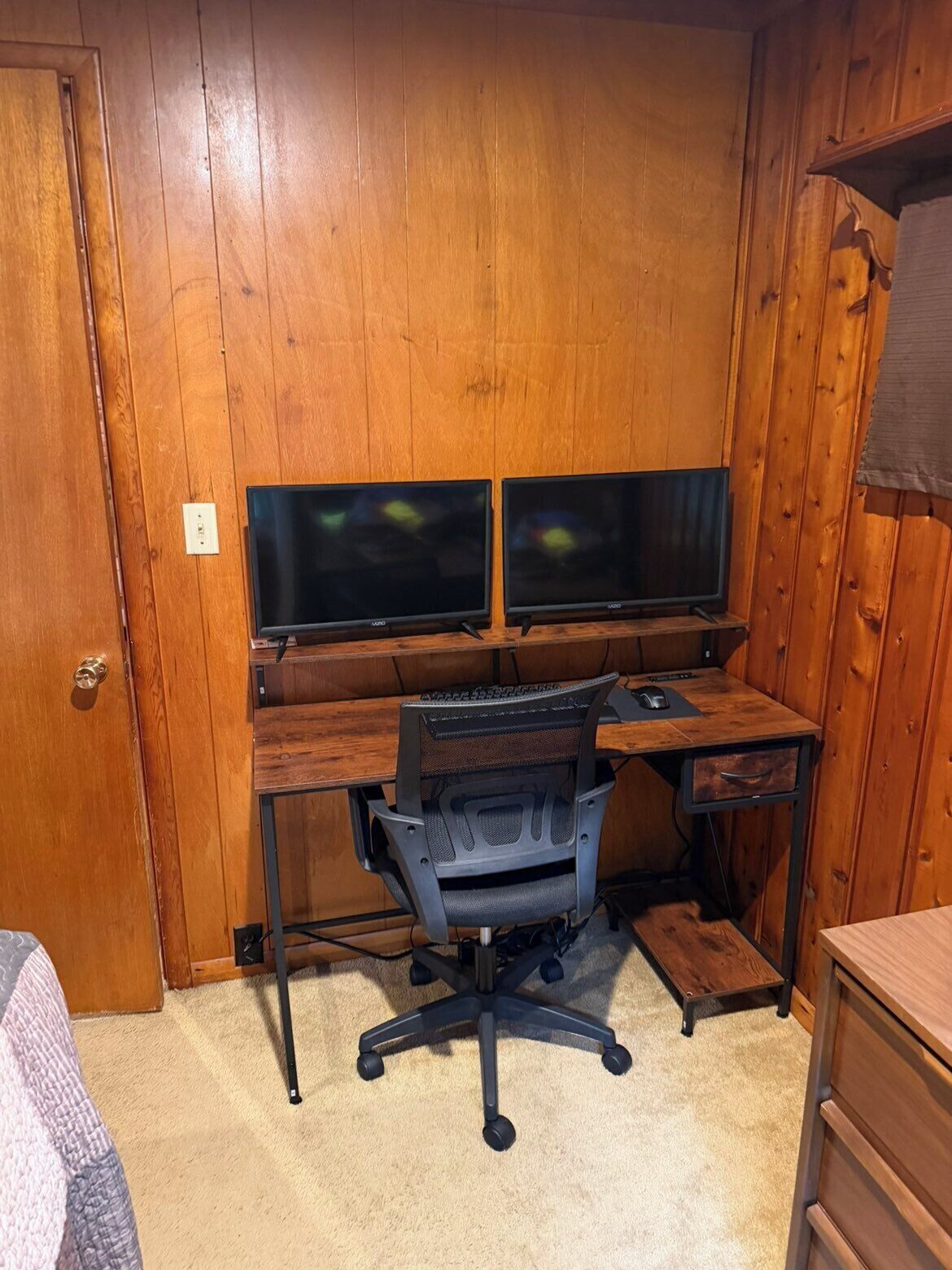 A room with wooden walls, a desk with two monitors, a chair, and a carpeted floor.