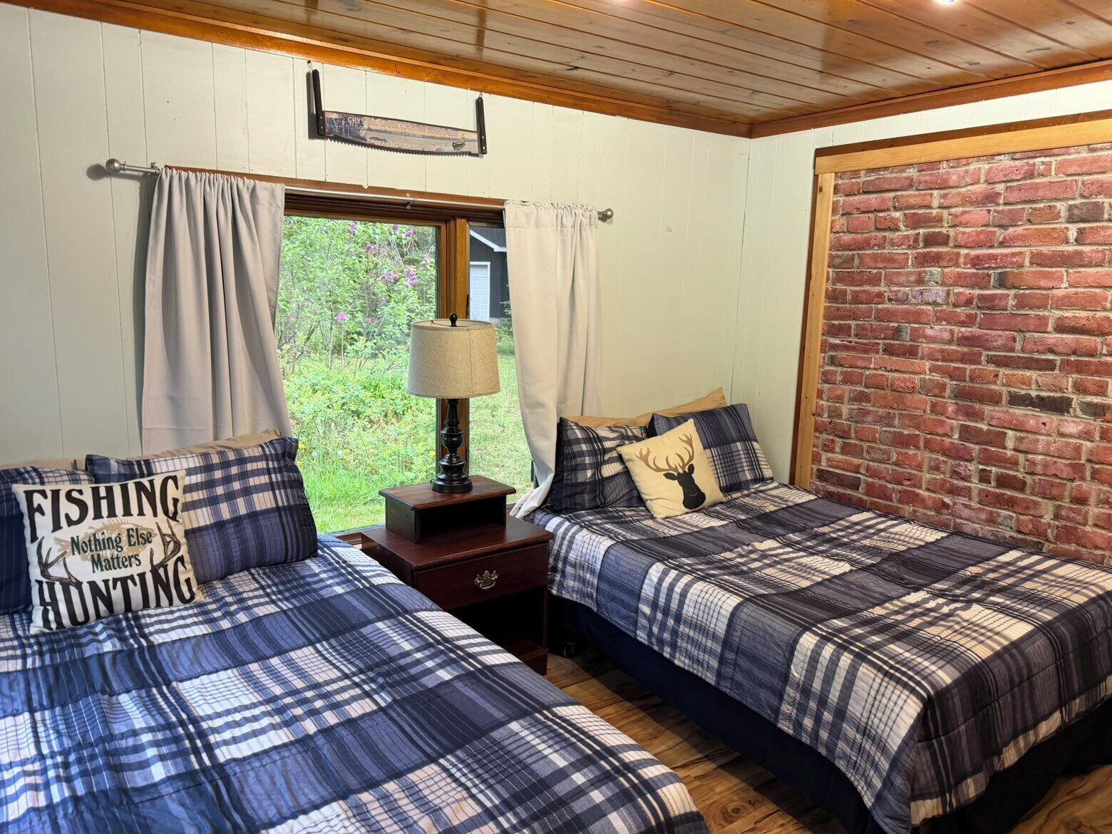 A room with two beds, a window with curtains, a lamp, and a brick wall.