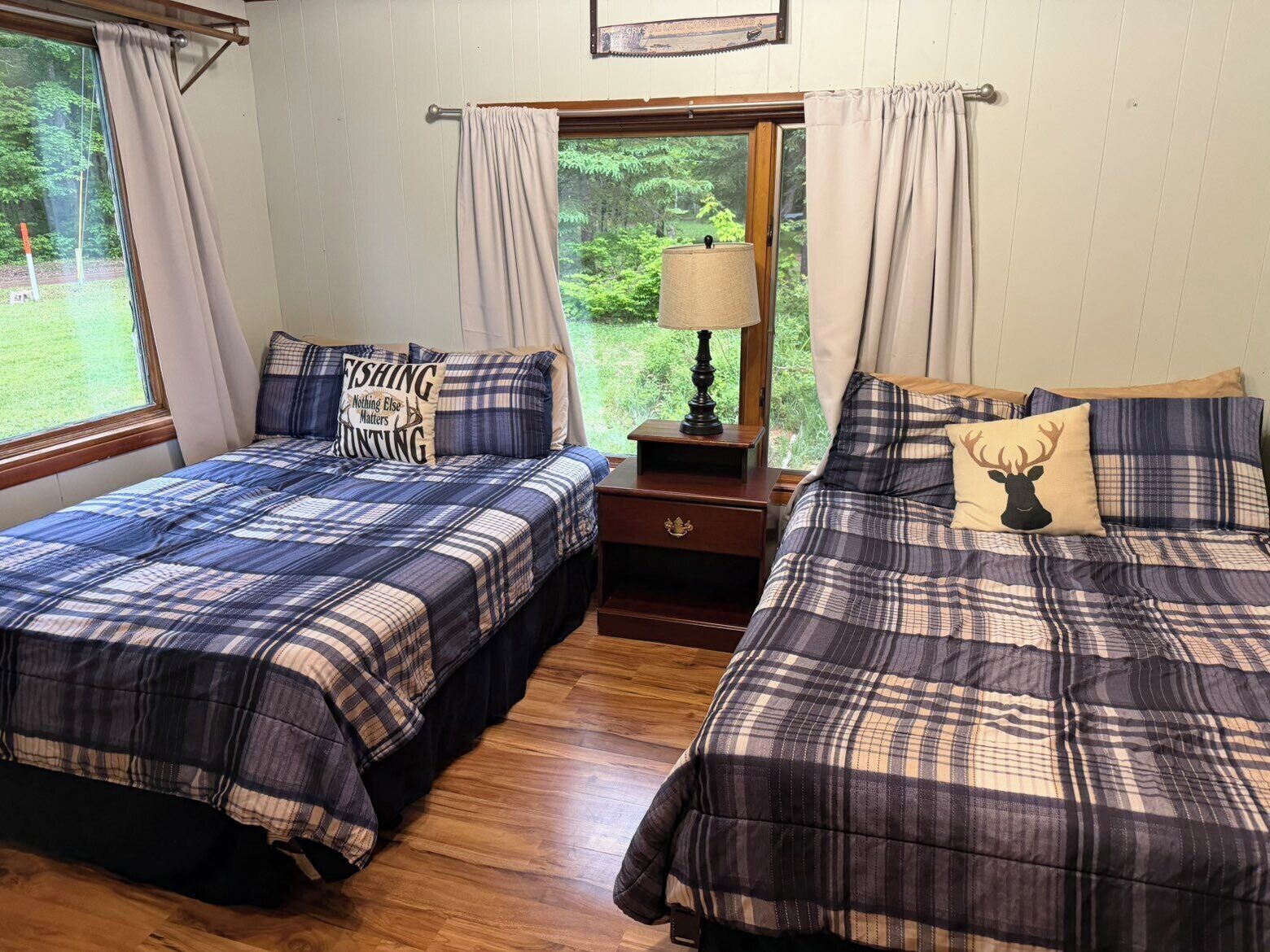Dos camas con ropa de cama escocesa, una mesita de noche con lámpara y un ventanal con cortinas.