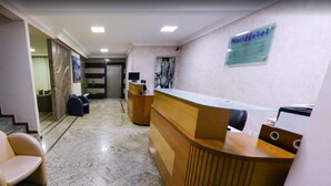 A hotel reception area with a reception desk, a seating area with chairs, and a staircase leading to another floor. - NortHotel (Barra De Sao Francisco)