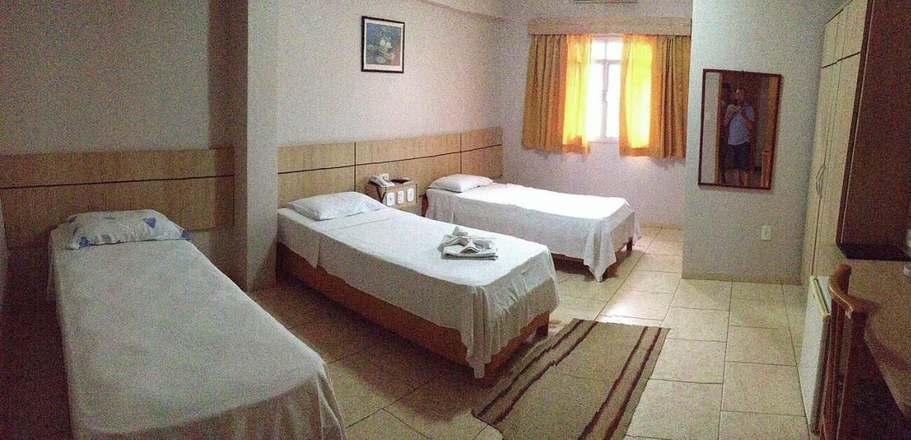 Quarto de hotel com duas camas de solteiro, uma mesinha de cabeceira com abajur, uma secretária pequena com cadeira, uma janela com cortinas e um espelho na parede.