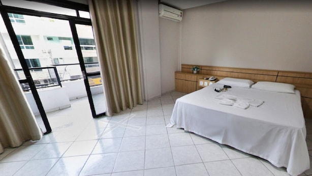 A hotel room with a bed, a bedside table with a phone and a small plant, and a view of a building through a glass door. - NortHotel (Barra De Sao Francisco)