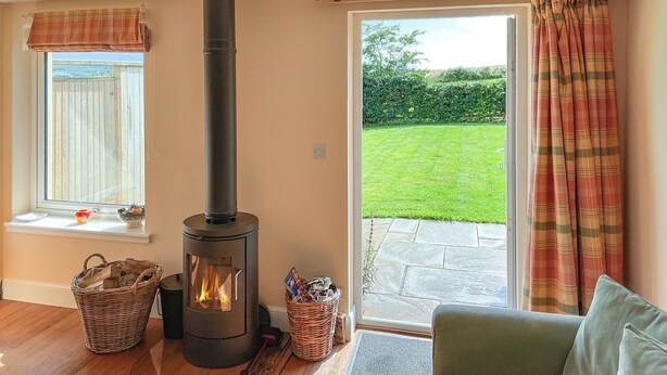 A cozy room with a wood-burning stove, a sofa, and a view of a garden through a sliding glass door. - The Annex at Fernyrig (Coldstream)