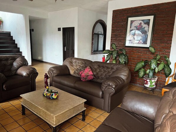 A living room with a brown leather sofa, a matching armchair, and a coffee table with a decorative item. There is a brick wall with a framed picture and a potted plant.