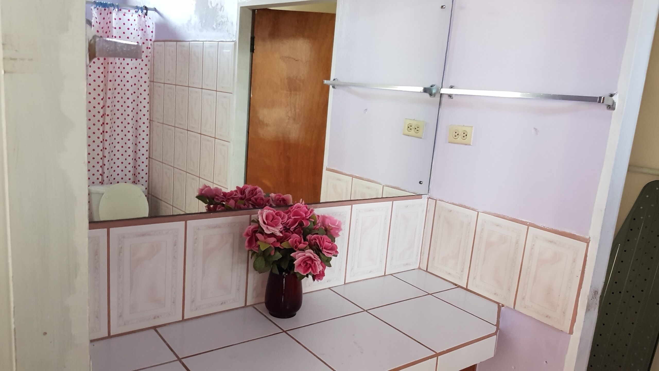 A bathroom with a mirror, a sink, a toilet, and a shower area. There is a vase with pink flowers on the sink counter.