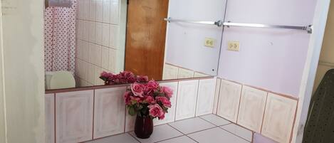 A bathroom with a mirror, a sink, a toilet, and a shower area. There is a vase with pink flowers on the sink counter.