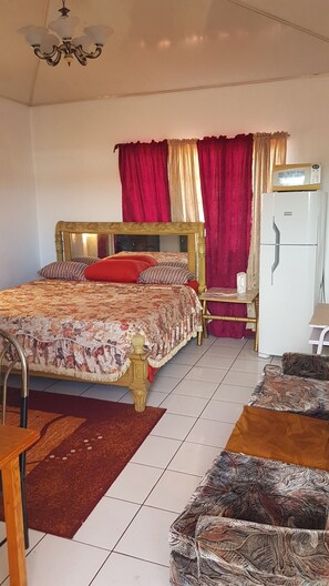 A bedroom with a bed, a bedside table, a chair, a refrigerator, and a chandelier. - Intercity Accommodations (St. Joseph)