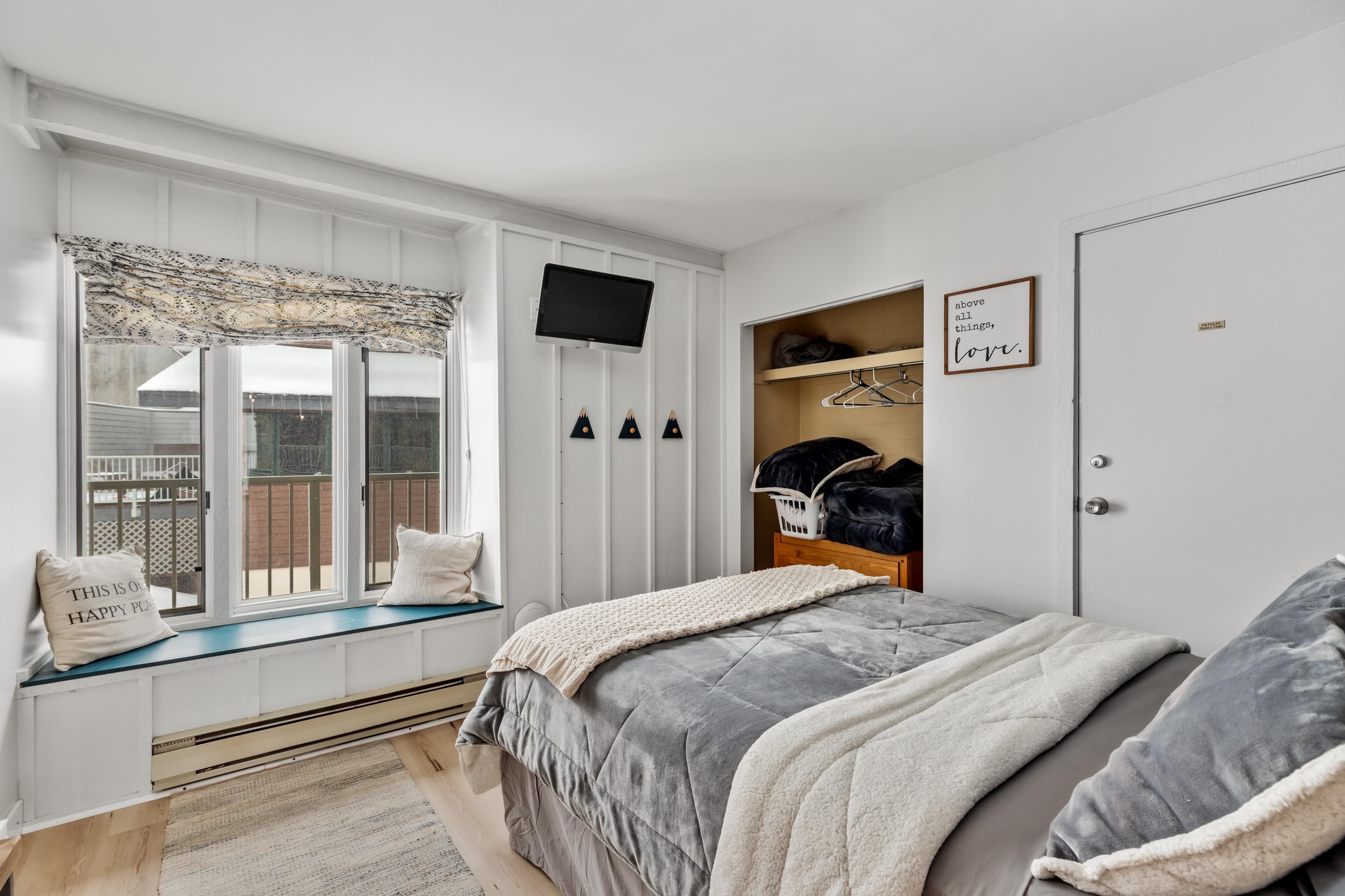 A bedroom with a bed, a window seat, a TV, a wardrobe, and a wall with a motivational quote.