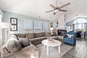 A living room with a sectional sofa, a round coffee table, and a blue armchair. There is a fireplace, a ceiling fan, and a dining area with a table and chairs. - Sound Of Dreams: Awesome Sound & Golf Views, Community Oceanfront Pool (Nags Head)