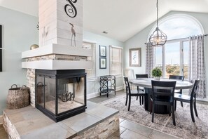 A dining area with a stone fireplace, a round table, and chairs. There are wicker baskets and a hanging light fixture. - Sound Of Dreams: Awesome Sound & Golf Views, Community Oceanfront Pool (Nags Head)