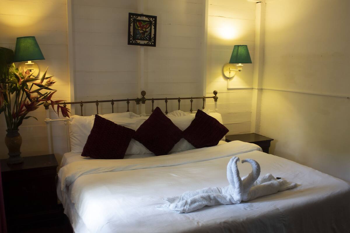 A neatly made bed with a swan-shaped towel, two green wall lamps, and a framed artwork above the bed.