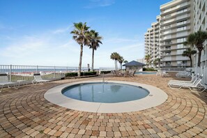 A pool area with lounge chairs, palm trees, and a building with balconies. - Windward Pointe 706 by Liquid Life (Orange Beach)