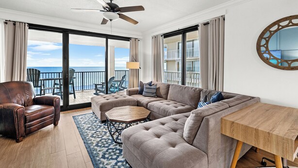 A spacious living room with a large sectional sofa, a wooden coffee table, and a leather armchair. A ceiling fan and a large window with a view of the ocean are visible.