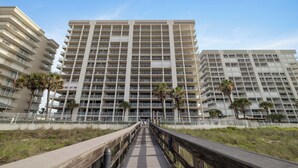 A multi-story hotel with a central walkway leading to the entrance, surrounded by palm trees and a white fence.