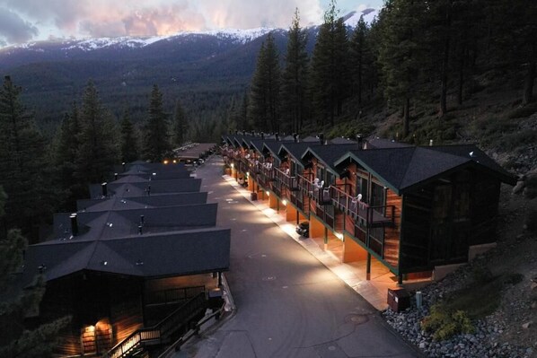 A row of log cabin-style lodges with a central walkway, surrounded by forested mountains.