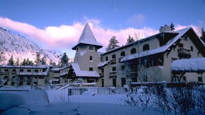 En snedækket bygning med et markant tårn, flere balkoner og et bjergrigt landskab i baggrunden.