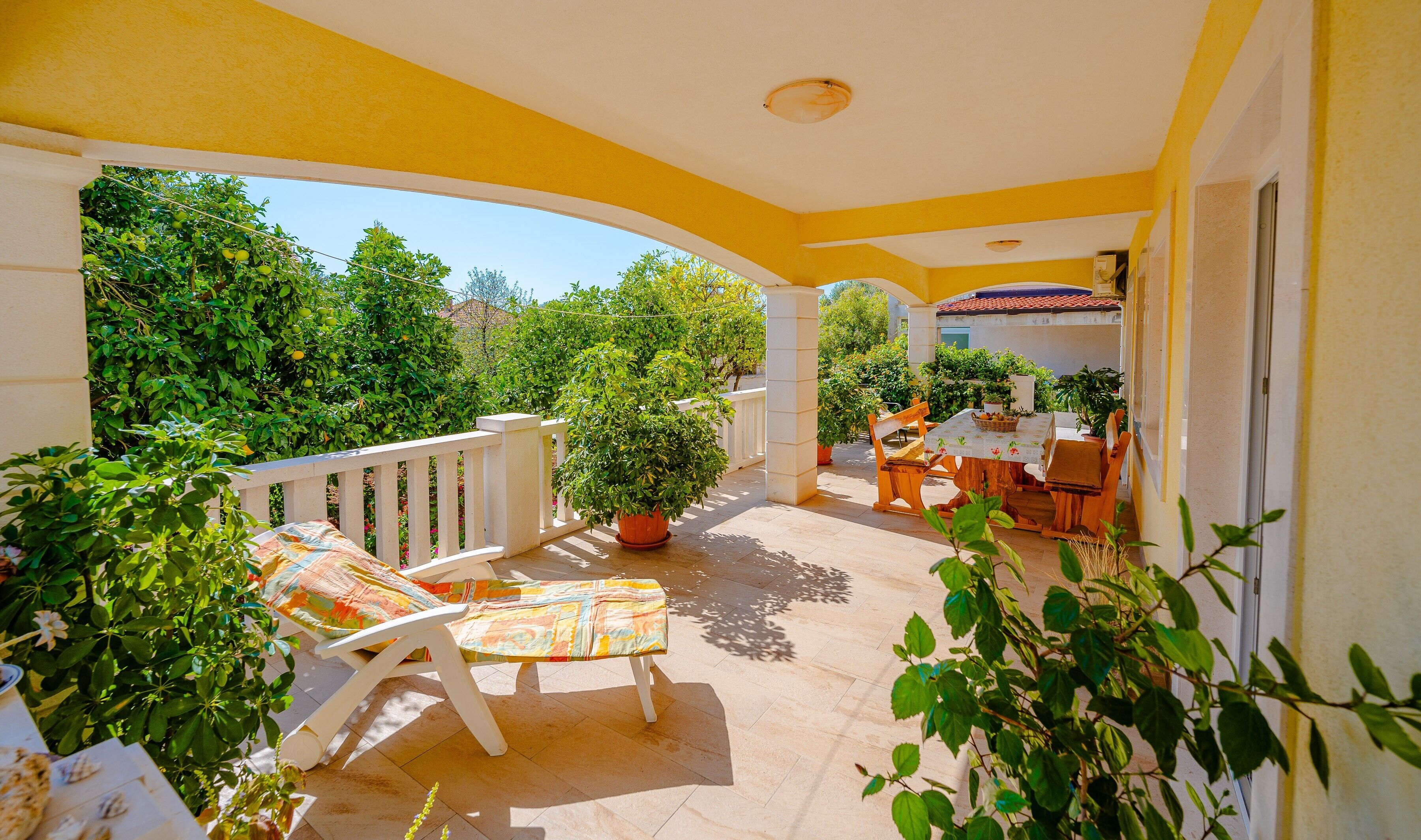 Eine überdachte Terrasse im Freien mit Tisch und Stühlen, umgeben von Topfpflanzen und Blick ins Grüne.