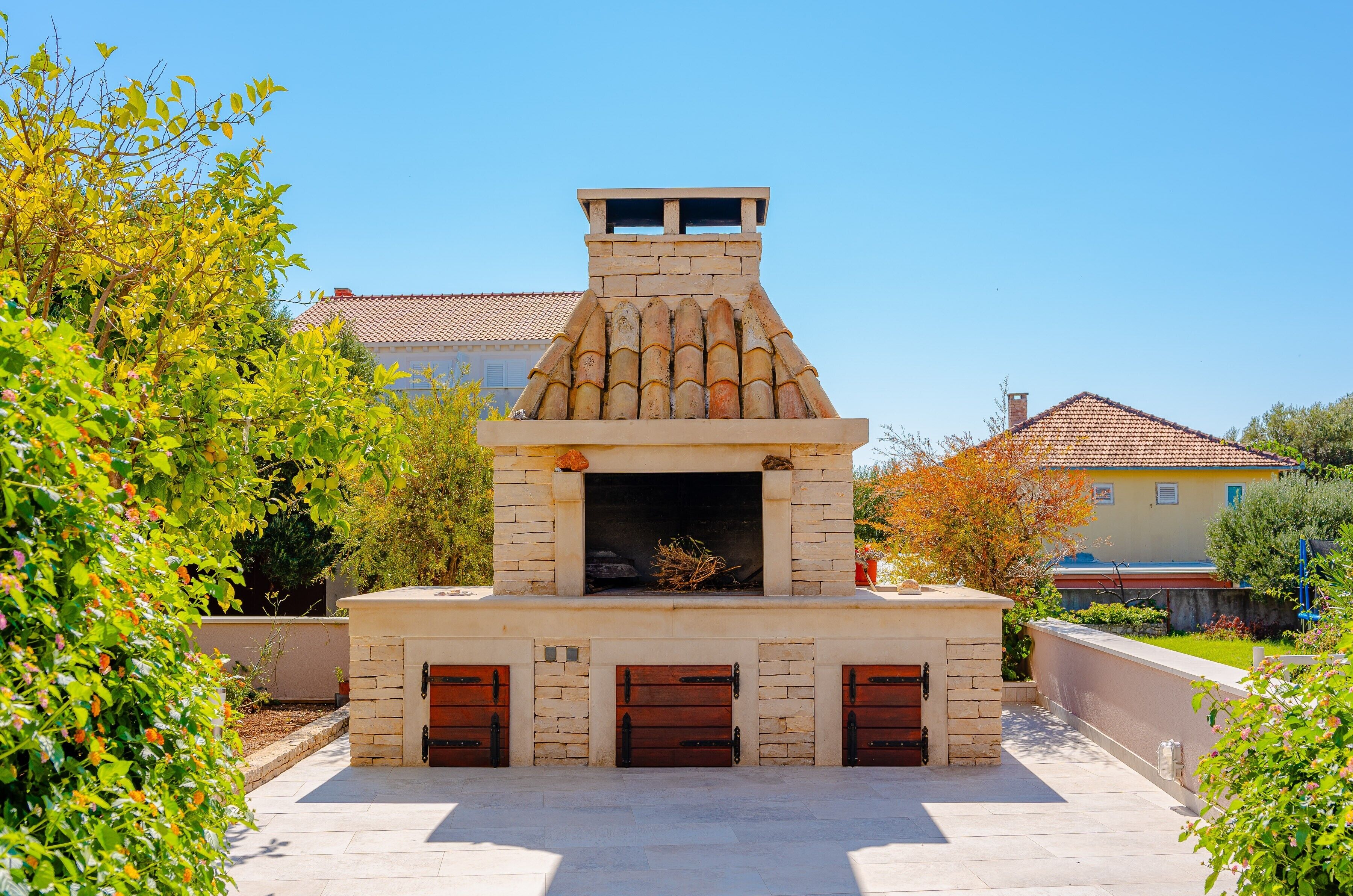A stone-built outdoor kitchen with a chimney, surrounded by greenery and a tiled roof in the background.