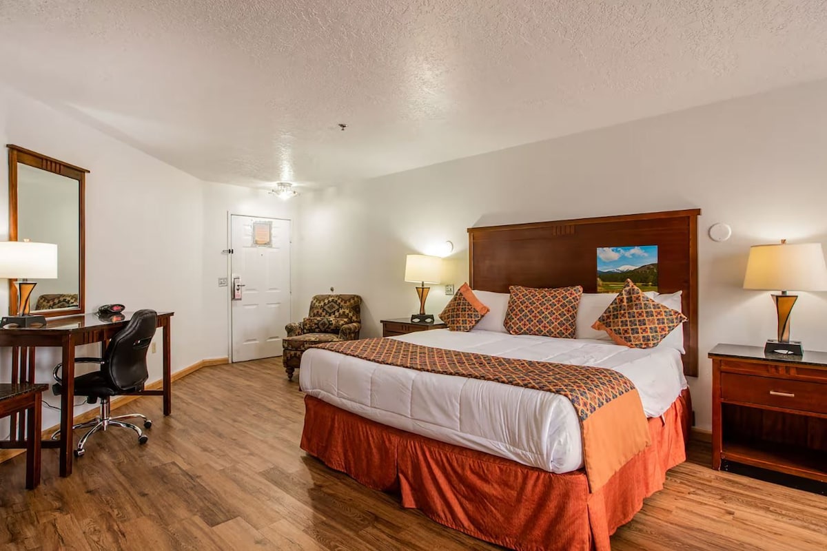 A hotel room with a large bed, a desk with a chair, a door, and a nightstand with a lamp.
