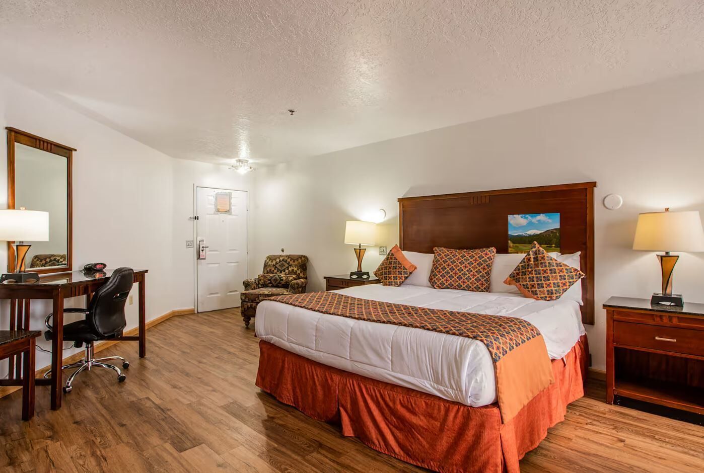 A hotel room with a large bed, a desk with a chair, a door, and a nightstand with a lamp.