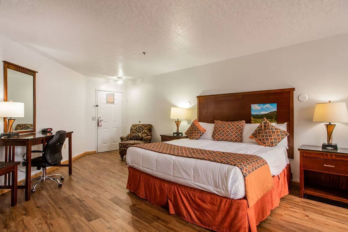 A hotel room with a large bed, a desk with a chair, a door, and a nightstand with a lamp.