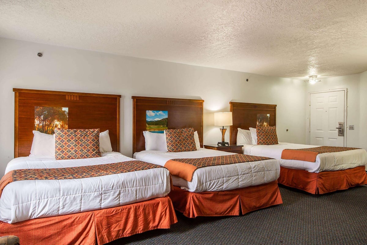 A hotel room with three beds, each with a wooden headboard, white bedding, and orange accents. There is a nightstand with a lamp and a painting on the wall.