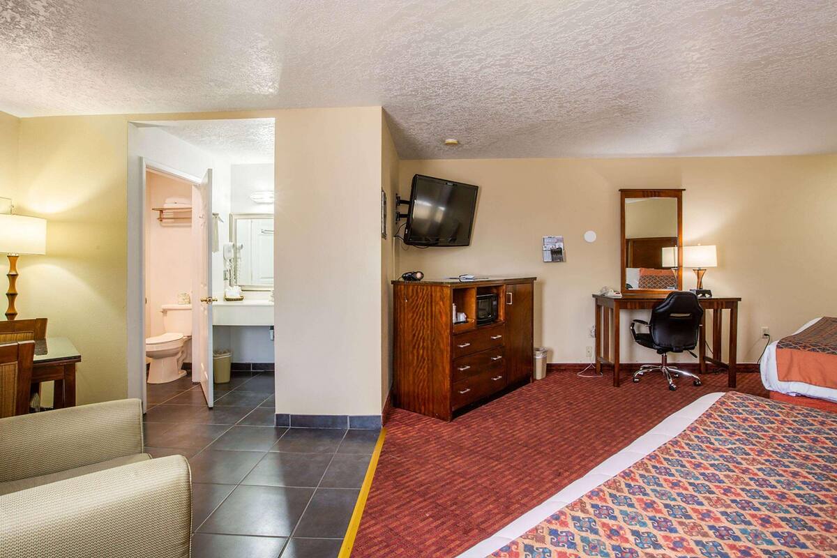 A hotel room with a bed, a desk with a chair, a television, a dresser, and a bathroom visible through an open door.