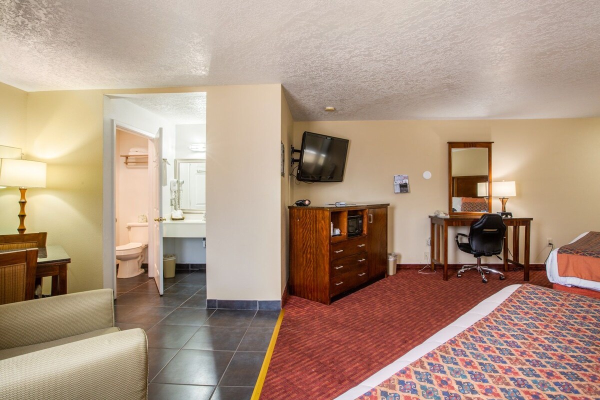 A hotel room with a bed, a desk with a chair, a television, a mirror, and a bathroom visible through an open door.