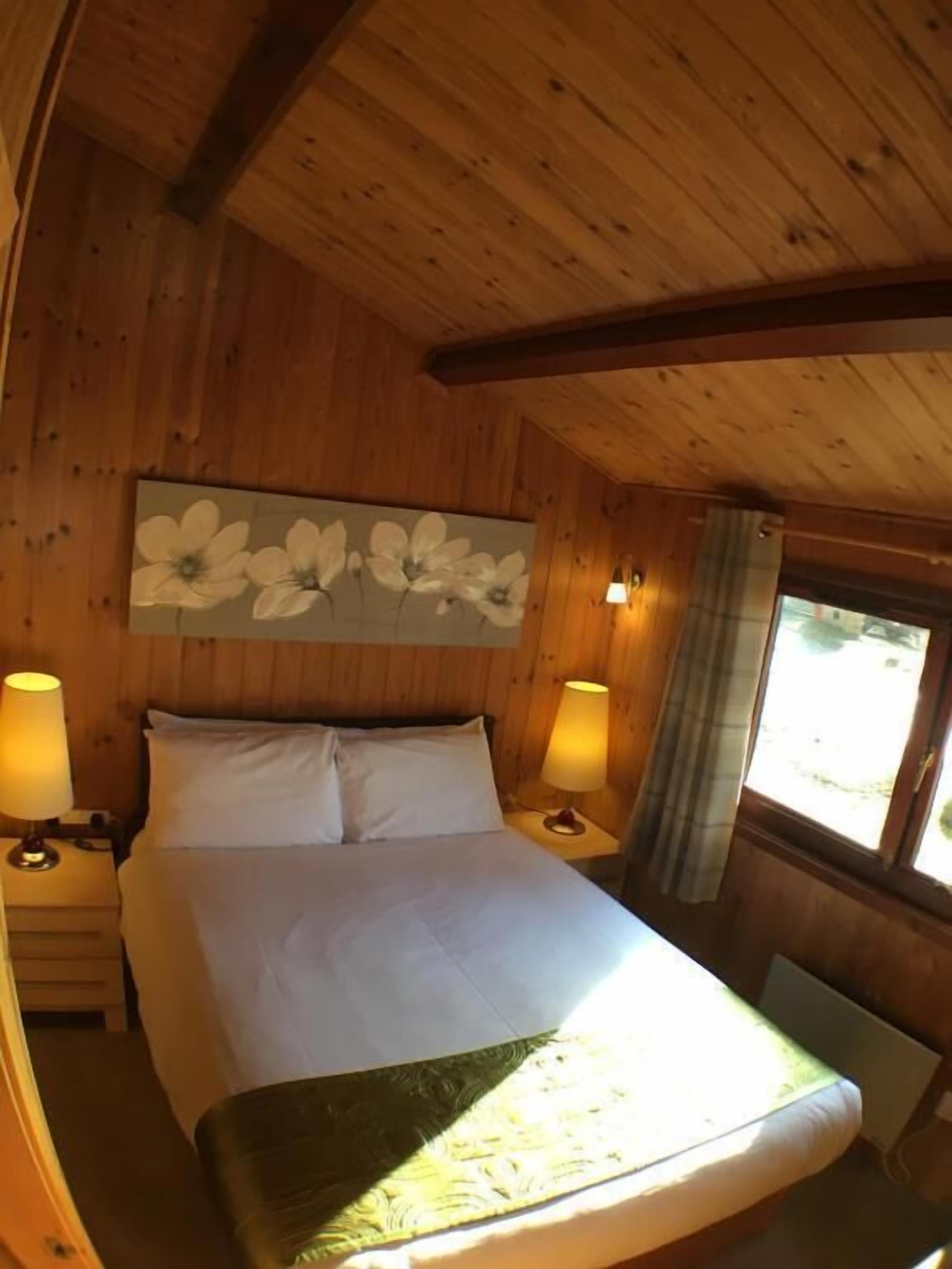 A wooden cabin room with a bed, two bedside lamps, a window with curtains, and a floral painting on the wall.