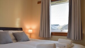 Un lit bien fait, avec du linge de lit et des serviettes blanches, une lampe de chevet et une fenêtre avec des rideaux.