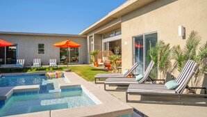 Een modern zwembadterras met ligbedden en parasols, omgeven door een verzorgd gazon en een gebouw met grote ramen.