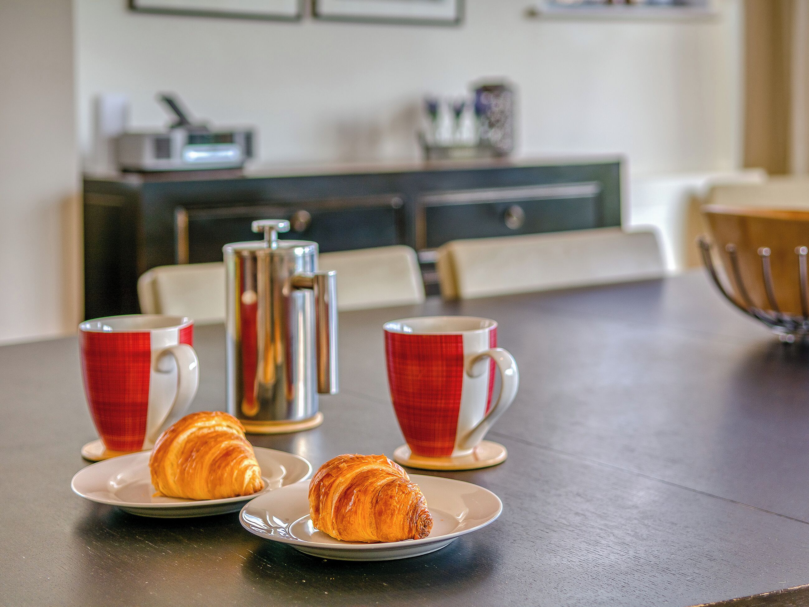 Una mesa de comedor con croissants y tazas rojas, una cafetera y un armario al fondo.