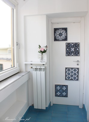 Une porte blanche ornée de carreaux décoratifs, un radiateur et une fenêtre donnant sur les bâtiments environnants.