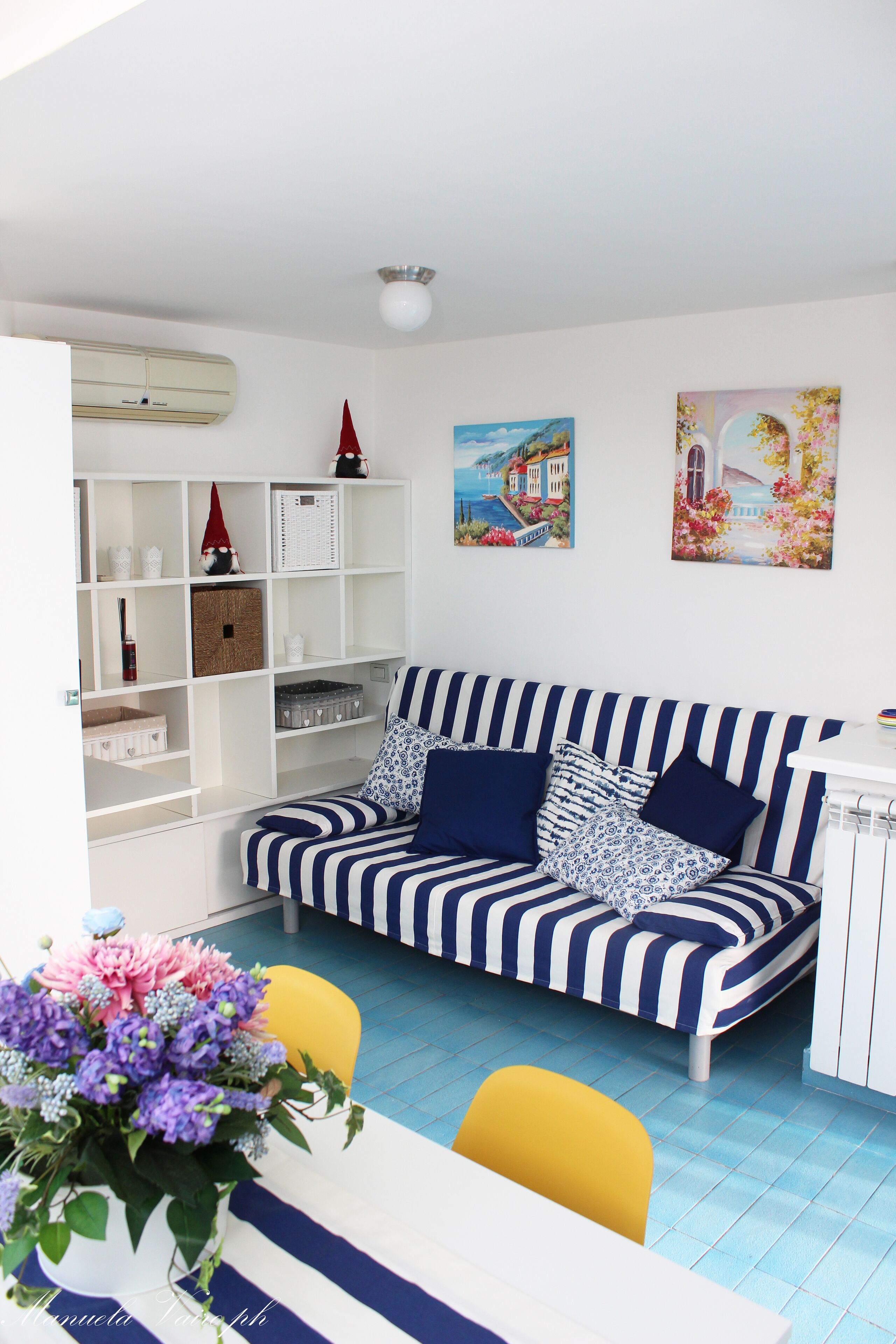 A living room with a striped sofa, a dining table, and a yellow chair. There is a white shelving unit with various items and two paintings on the wall.