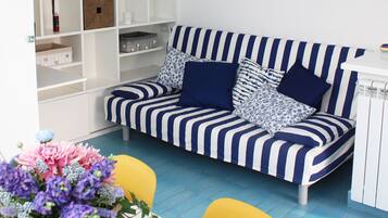 Ein Wohnzimmer mit einem gestreiften Sofa, einem Esstisch und einem gelben Sessel. Eine weiße Regaleinrichtung mit diversen Gegenständen und zwei Bildern an der Wand.