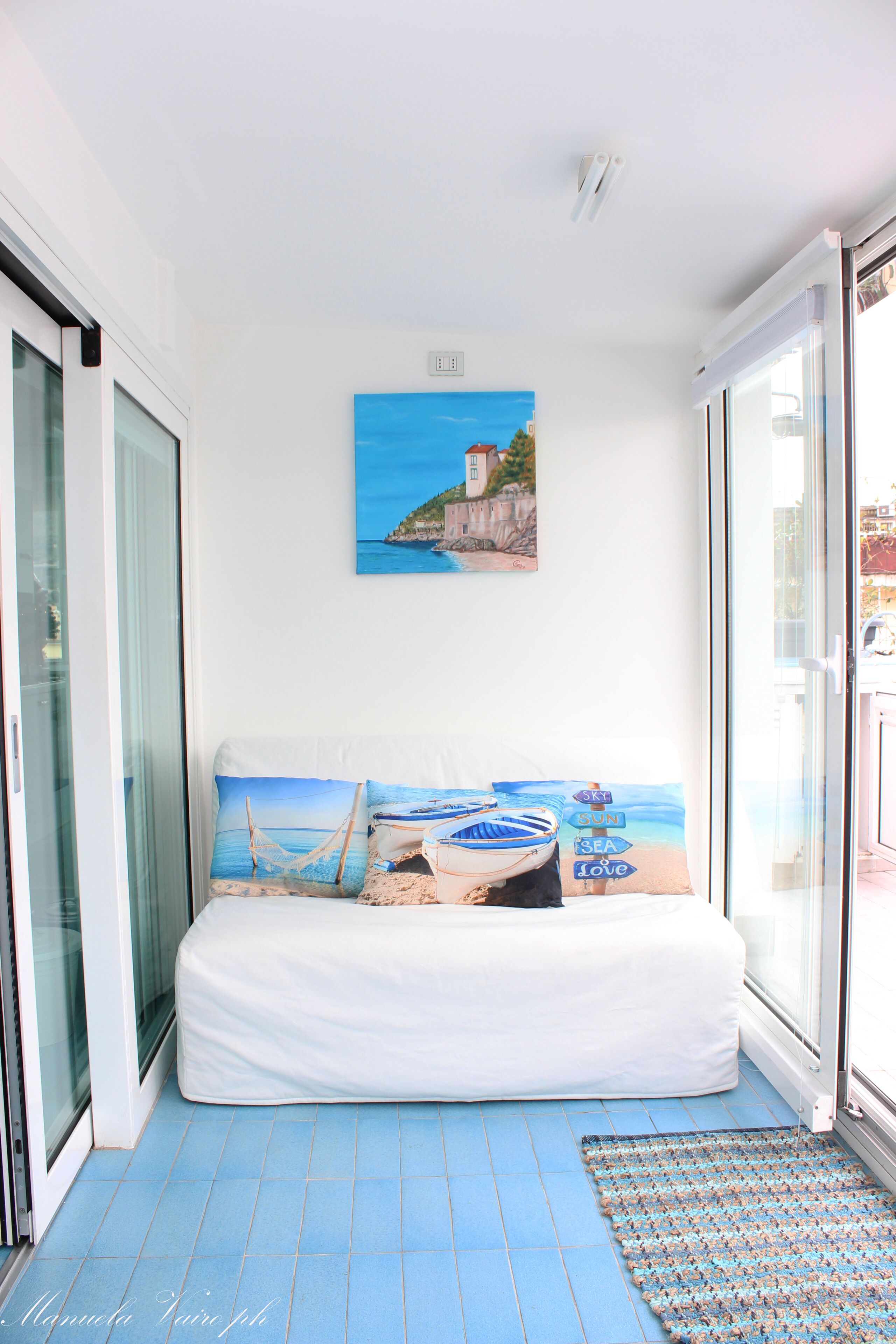 A bedroom with a bed, a painting of a coastal scene, and a view through a glass door.