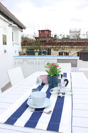Une table à manger dressée avec une nappe rayée bleue et blanche, une tasse blanche, un pot rose contenant des fleurs et une bouilloire argentée.