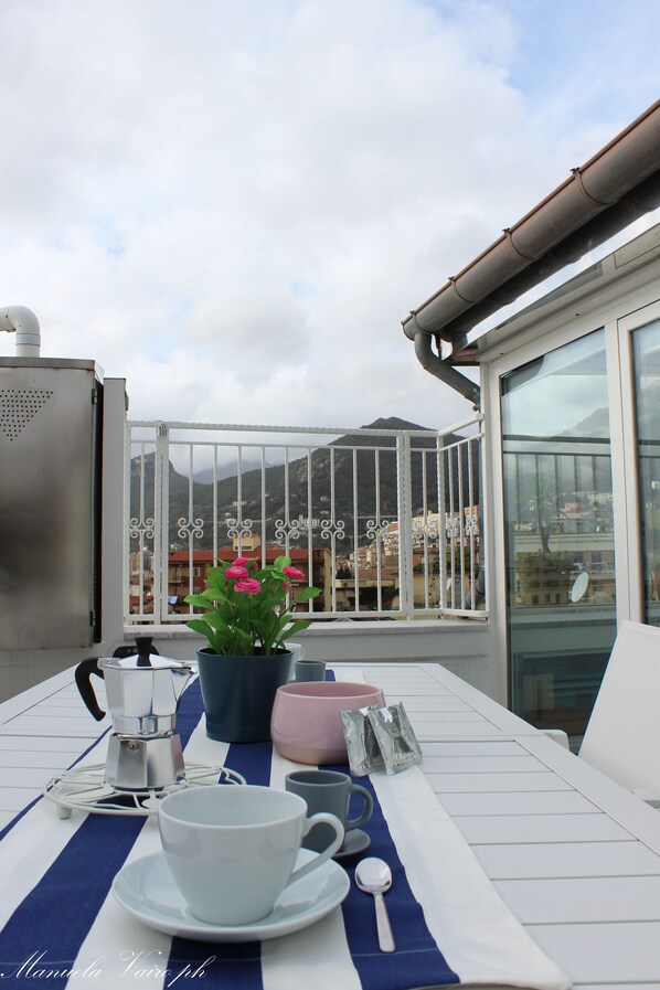 Een balkon met een tafel gedekt voor twee personen, een koffiezetapparaat en een bloeiende plant in een pot.