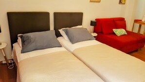 Une chambre d’hôtel avec deux lits, chacun doté d’une tête de lit et d’oreillers. On y trouve une chaise rouge et une petite table avec une lampe.