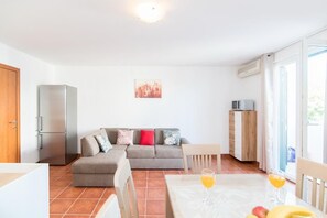 A modern living room with a sofa, a dining table with fruit and drinks, a refrigerator, and a cityscape painting.