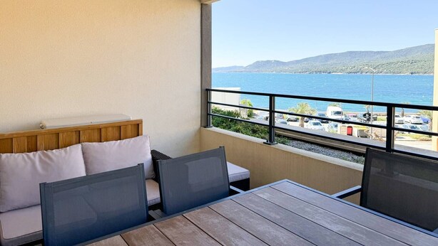 A balcony with a wooden table and chairs, a cushioned bench, and a view of the sea and distant hills. - Fronte di Mare 2 bedroom town center sea view (PROPRIANO)