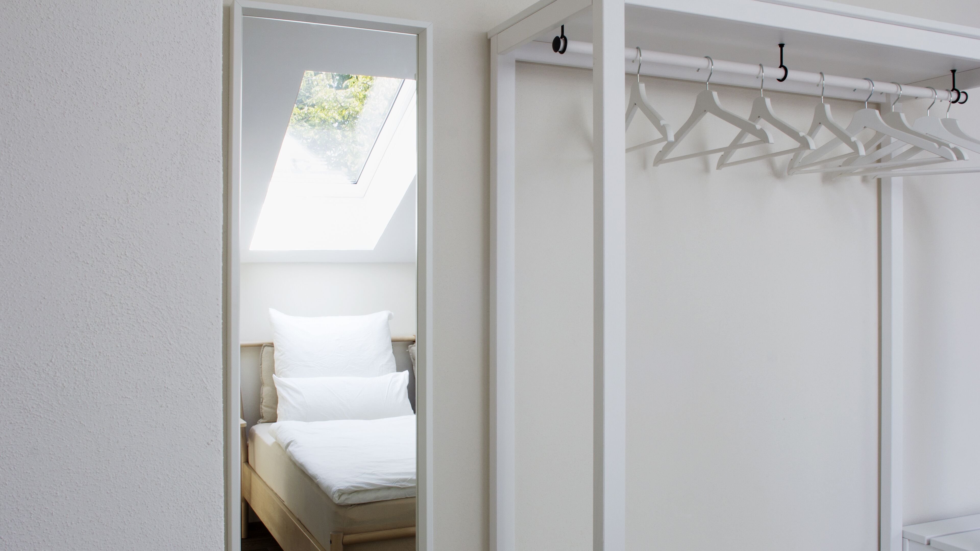 Un dormitorio con cama, tragaluz y armario blanco con perchas.