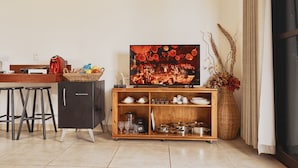 Una cocina con televisión, taburetes, refrigerador, un armario de madera con vajilla y un florero con flores secas.