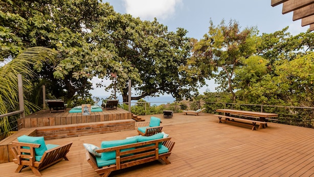 Una terraza de madera con mobiliario exterior, que incluye sillas y un banco, rodeada de abundante follaje y árboles.