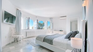 A modern bedroom with a large bed, a bedside table, a chair, and a TV mounted on the wall. - Majestic Panorama Ocean View Villa Multicolor LED XXL Pool Ibiza Town (Can Furnet)