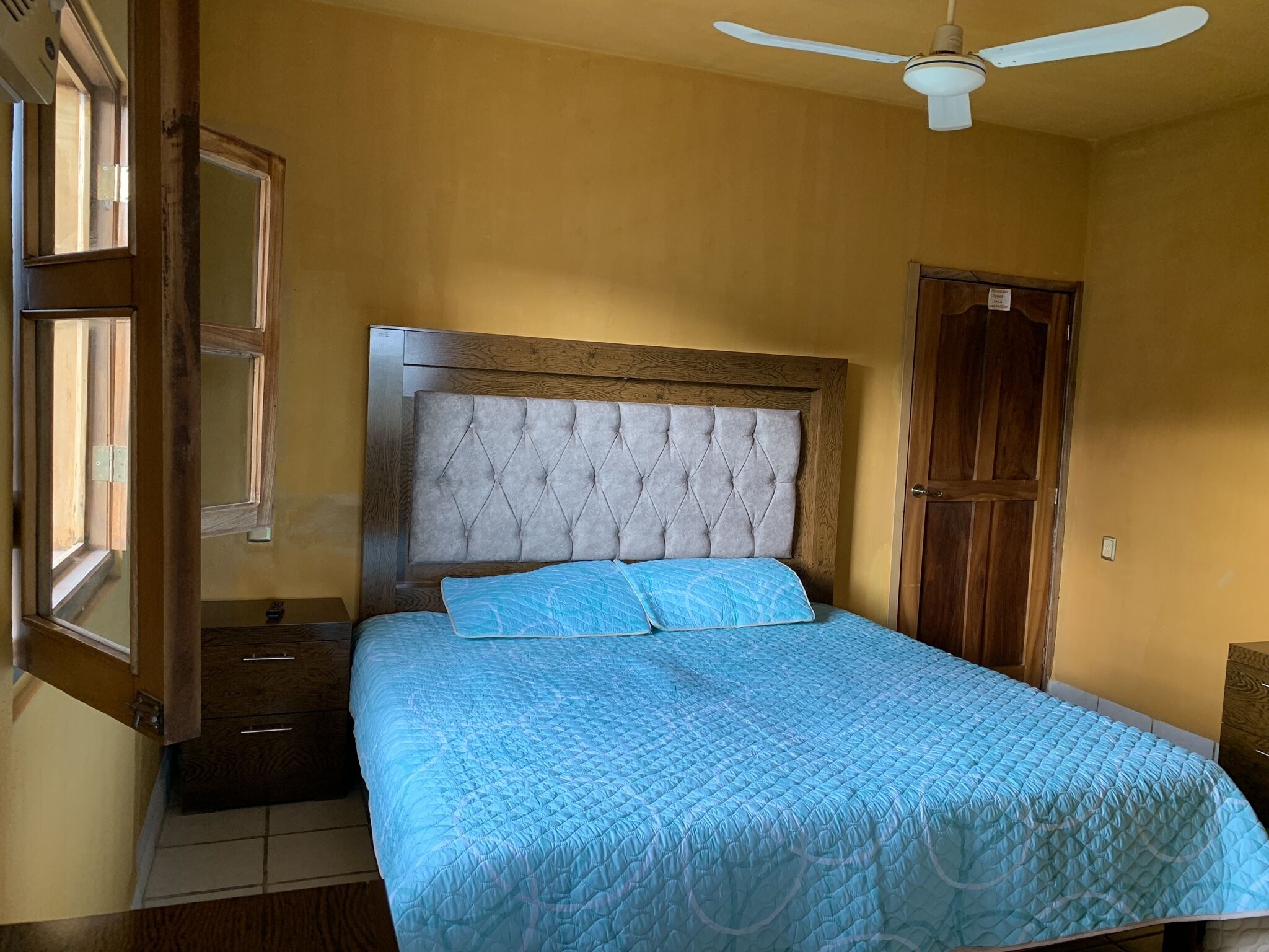 A bedroom with a wooden headboard, a bed with a blue quilt, a ceiling fan, and a wooden wardrobe.